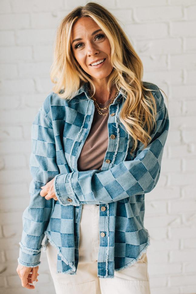 Woman wearing a blue plaid shirt over a brown top against a white brick wall.