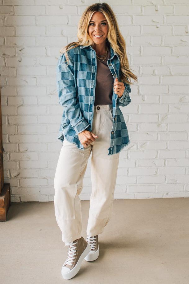 Woman wearing a blue plaid shirt, beige pants, and white sneakers against a white brick wall.