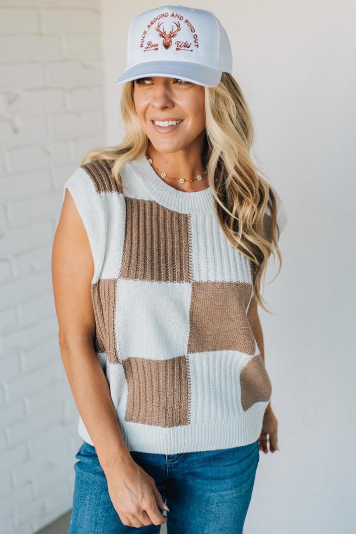 Woman wearing a checkered sleeveless sweater and cap with a logo against a white brick wall.