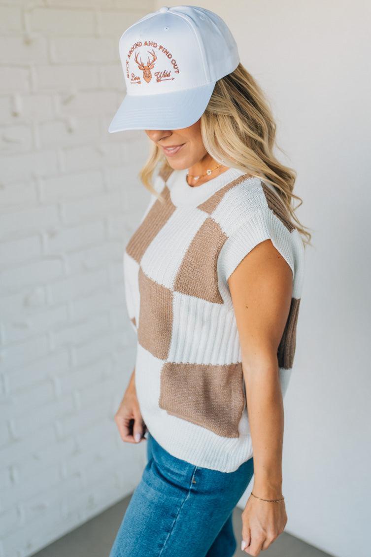 Woman wearing a patterned sleeveless sweater and cap against a white brick wall.