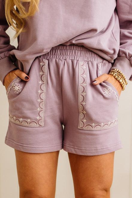 Person wearing lavender shorts with intricate white embroidery on a plain background