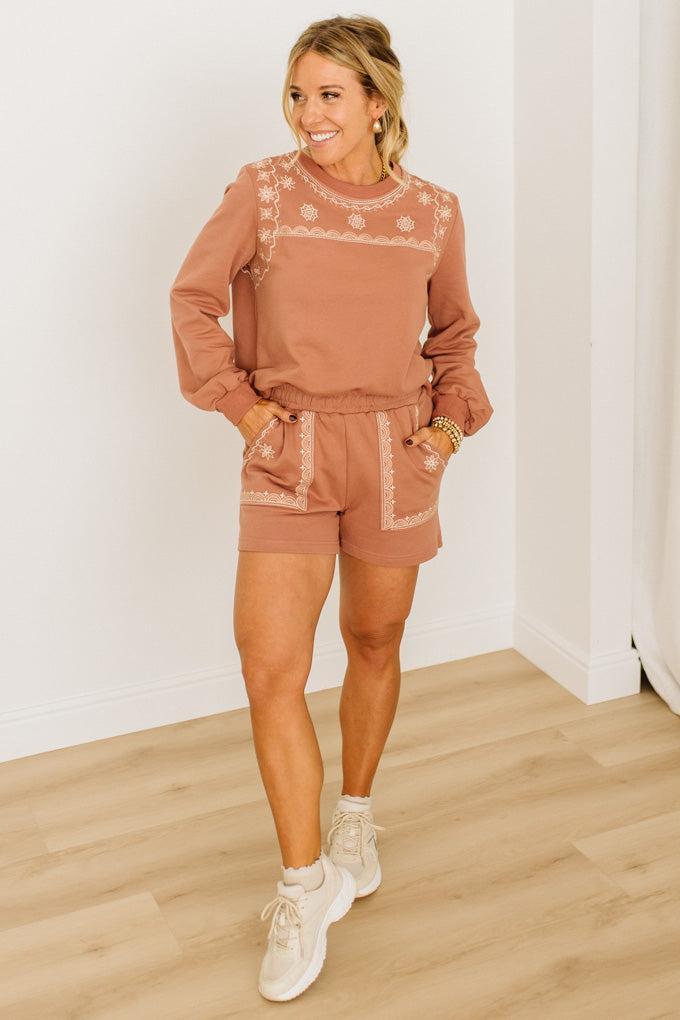 Woman wearing a brown embroidered outfit with white sneakers on a wooden floor.