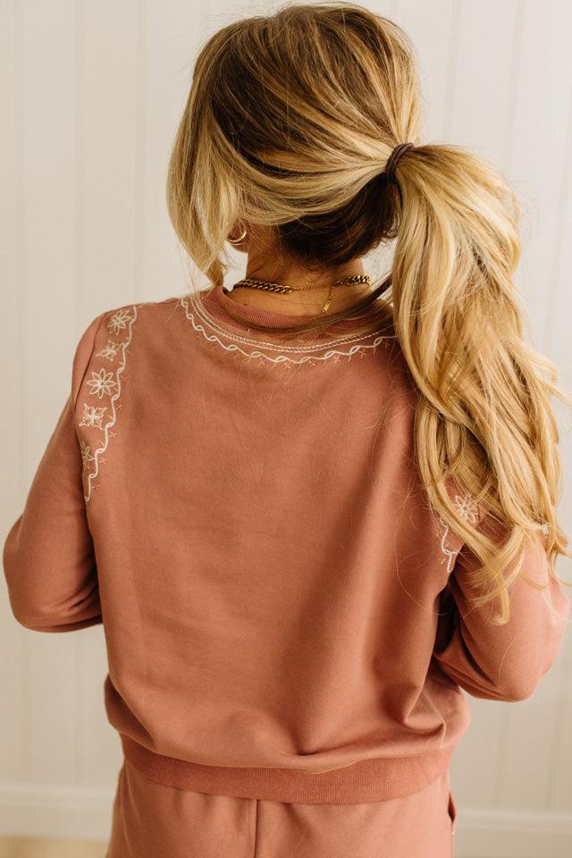 Person wearing a brown sweatshirt with embroidered details, standing against a neutral background.