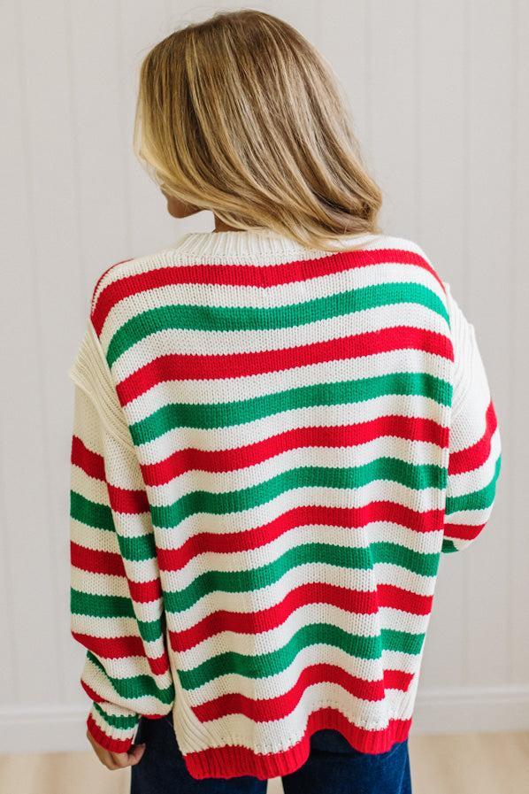 BLONDE WOMAN IN DARK JEANS STANDING IN A BRIGHTLY LIT STUDIO AND WEARING A CHRISTMAS CANDY STRIPE COZY KNIT PULLOVER IN CREAM GREEN AND RED.
