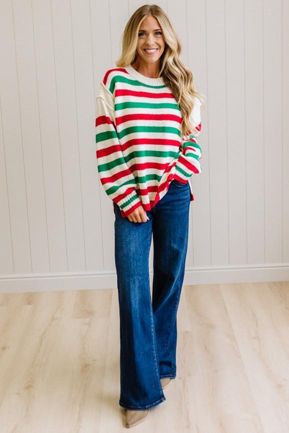BLONDE WOMAN IN DARK JEANS STANDING IN A BRIGHTLY LIT STUDIO AND WEARING A CHRISTMAS CANDY STRIPE COZY KNIT PULLOVER IN CREAM GREEN AND RED.