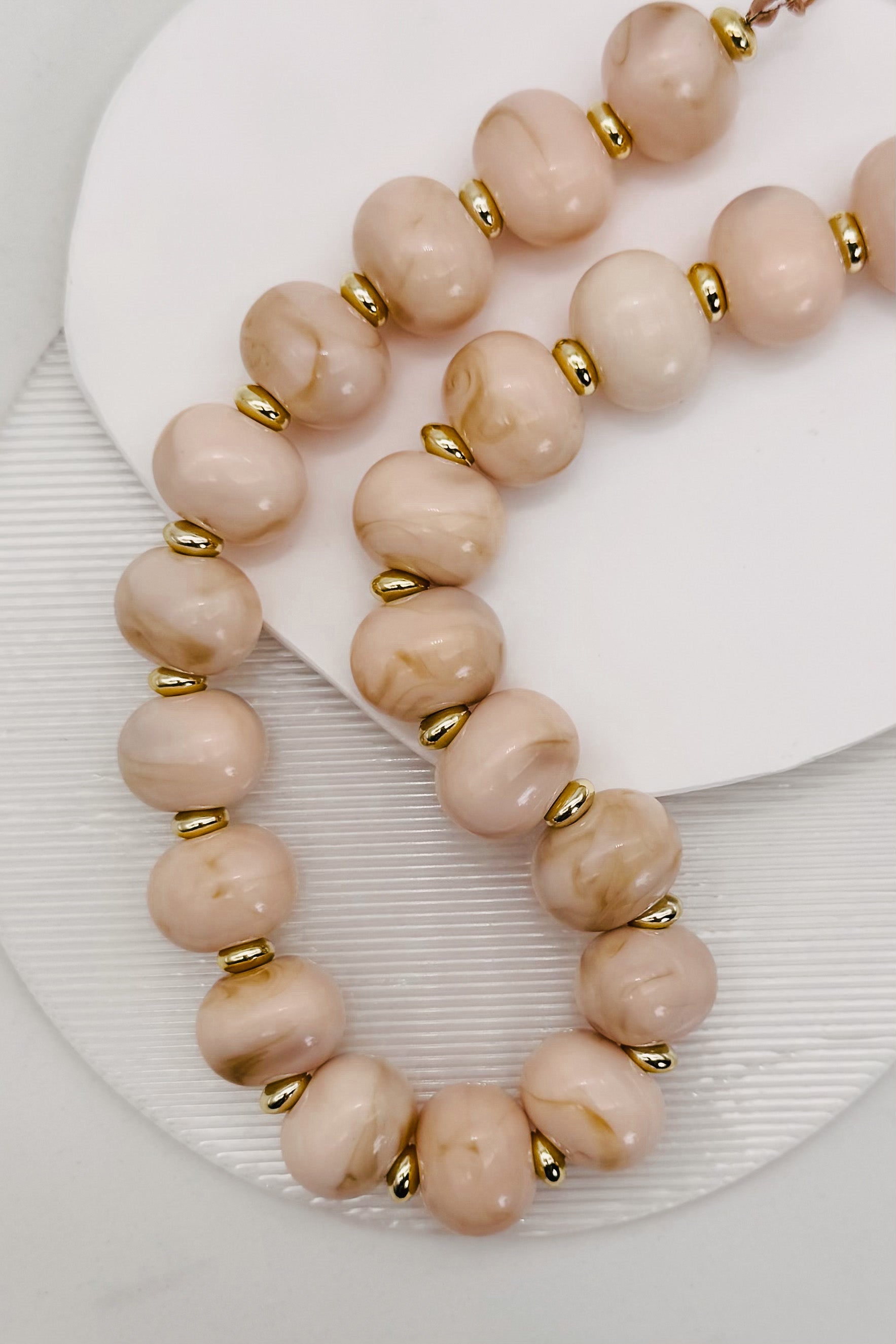 A close-up of a beaded necklace with large, round, dusty pink stone beads separated by small gold metal spacers, displayed on a white textured surface.