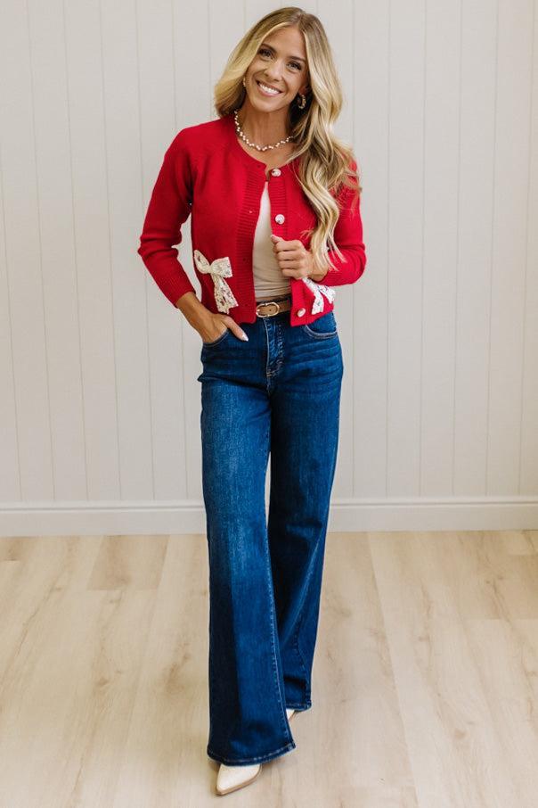 BLONDE WOMAN IN A LIGHT STUDIO WEARING DARK JEANS AND A LIGHT TOP WITH A RED CARDIGAN OVER TOP THAT HAS LARGE LACE BOWS ON THE SIDES.