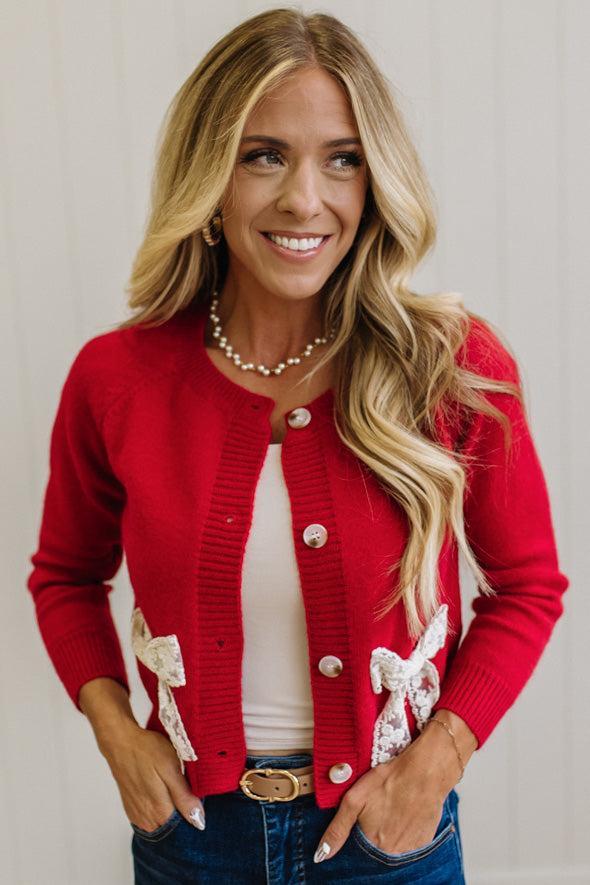 BLONDE WOMAN IN A LIGHT STUDIO WEARING DARK JEANS AND A LIGHT TOP WITH A RED CARDIGAN OVER TOP THAT HAS LARGE LACE BOWS ON THE SIDES.