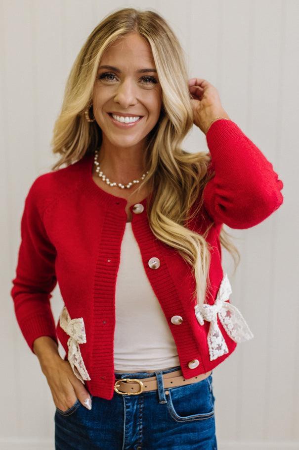 BLONDE WOMAN IN A LIGHT STUDIO WEARING DARK JEANS AND A LIGHT TOP WITH A RED CARDIGAN OVER TOP THAT HAS LARGE LACE BOWS ON THE SIDES.