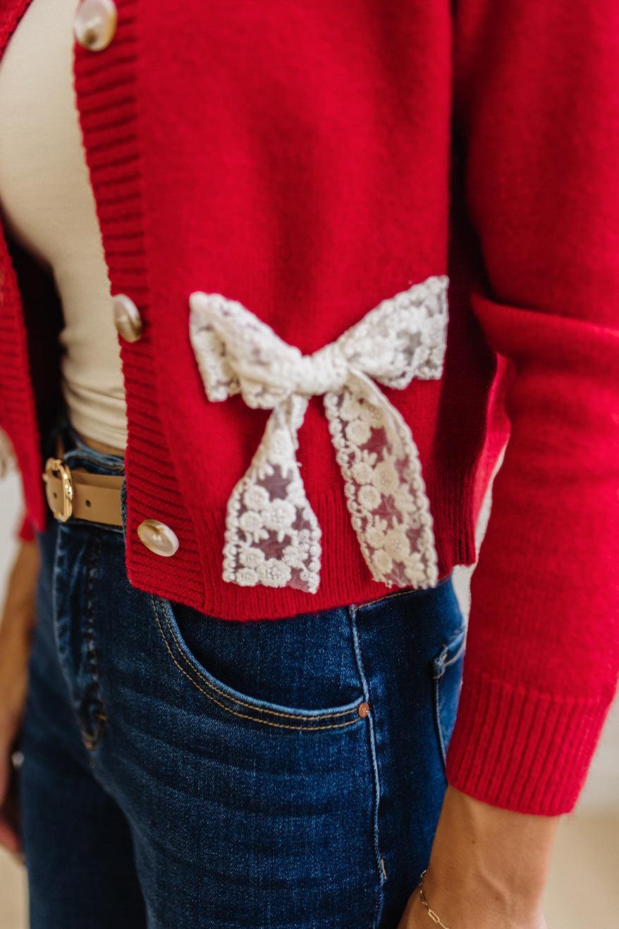 BLONDE WOMAN IN A LIGHT STUDIO WEARING DARK JEANS AND A LIGHT TOP WITH A RED CARDIGAN OVER TOP THAT HAS LARGE LACE BOWS ON THE SIDES.