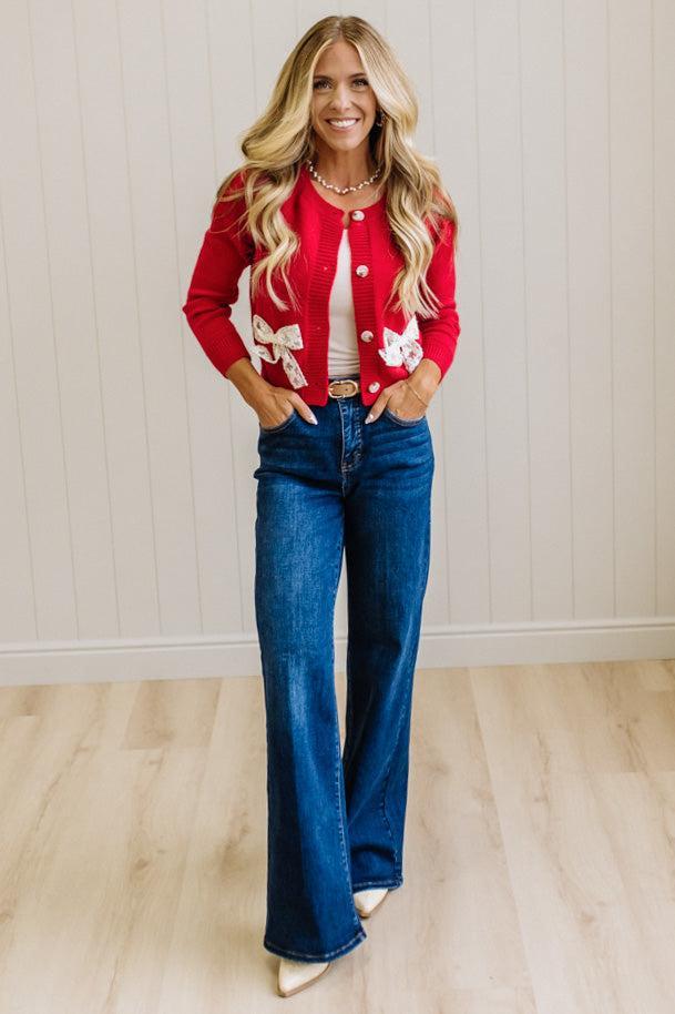 BLONDE WOMAN IN A LIGHT STUDIO WEARING DARK JEANS AND A LIGHT TOP WITH A RED CARDIGAN OVER TOP THAT HAS LARGE LACE BOWS ON THE SIDES.