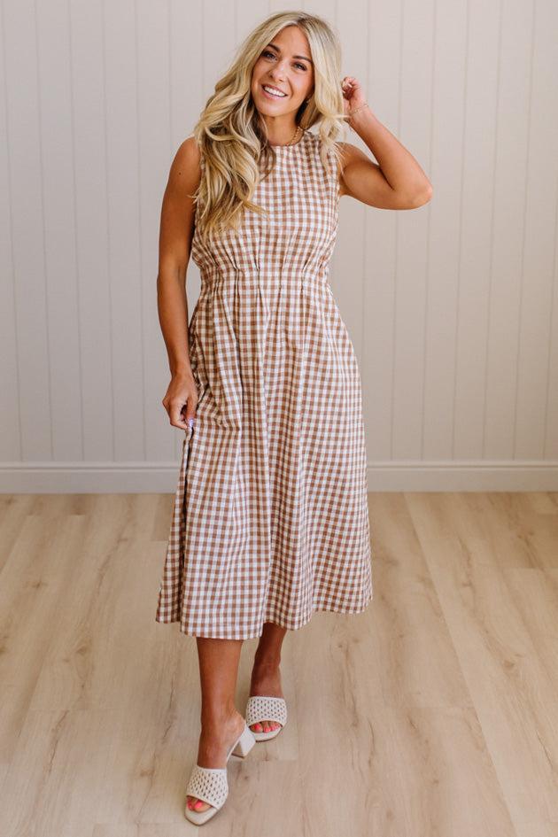 Woman wearing a checkered dress standing on a wooden floor with a white paneled wall in the background.