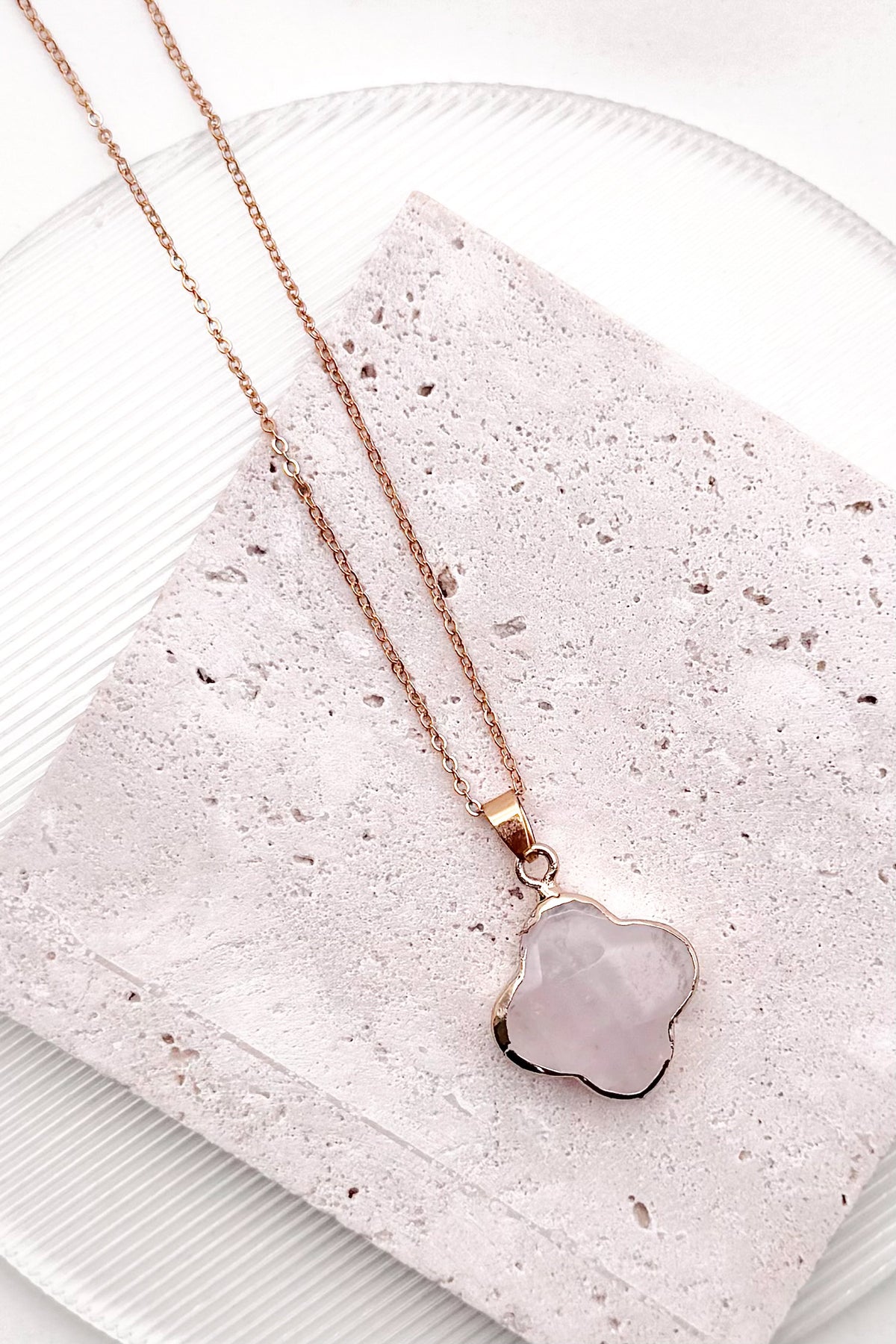Rose gold necklace with a polished, irregular-shaped pale pink stone pendant, displayed on a textured light pink stone tile over a white plate.