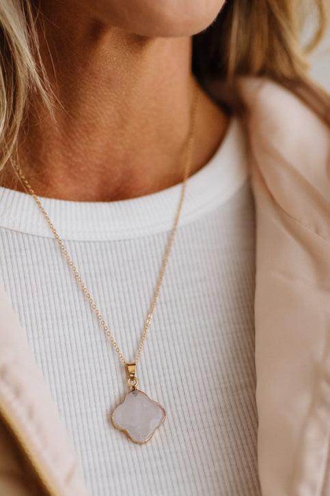 A close-up lifestyle shot of a woman wearing a delicate gold chain necklace with a light pinkish-white clover-shaped pendant, styled over a white ribbed top and a beige jacket.