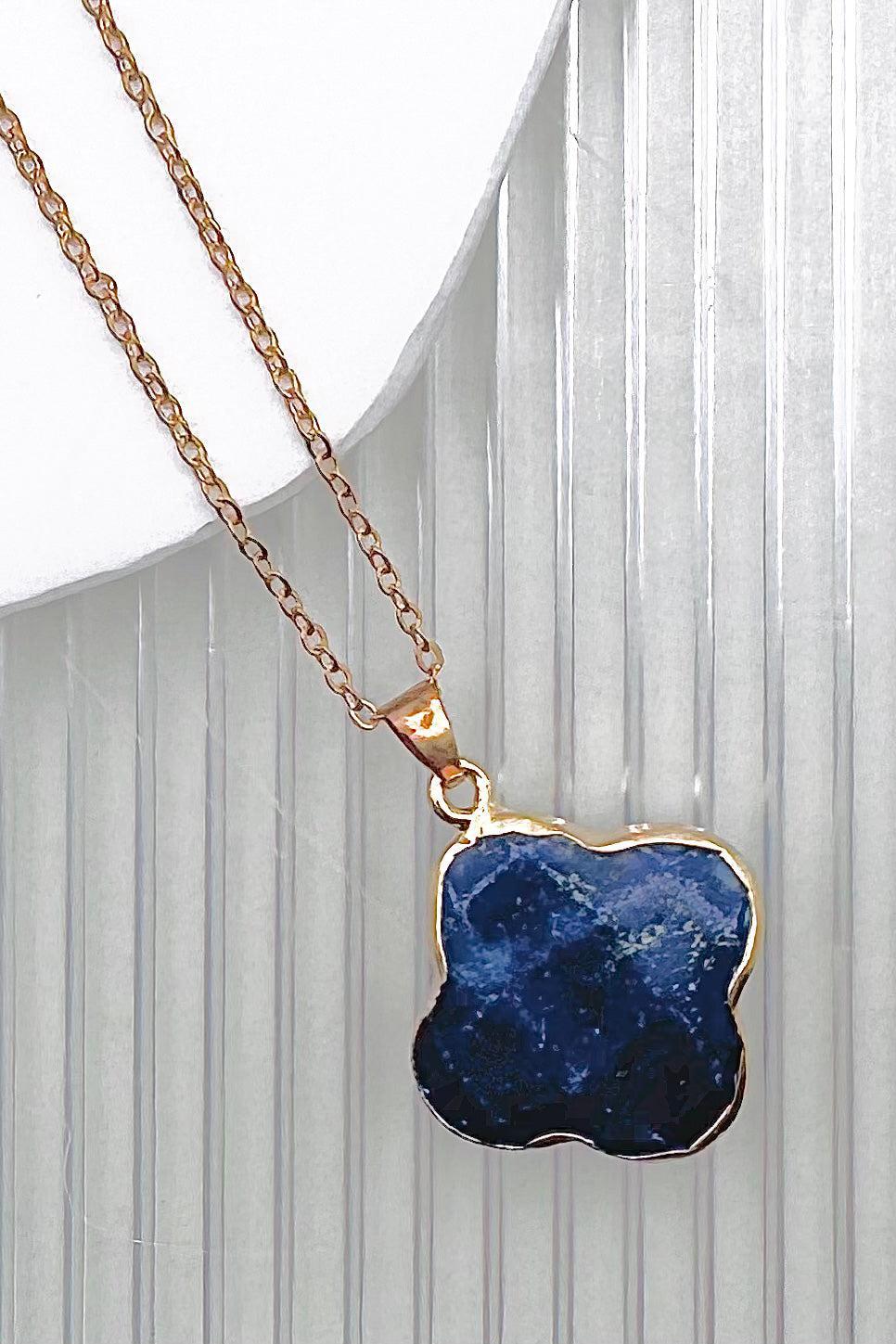A gold necklace with a wavy clover-shaped pendant made of deep blue lapis lazuli stone, displayed against a white background with vertical gray ribbed lines.
