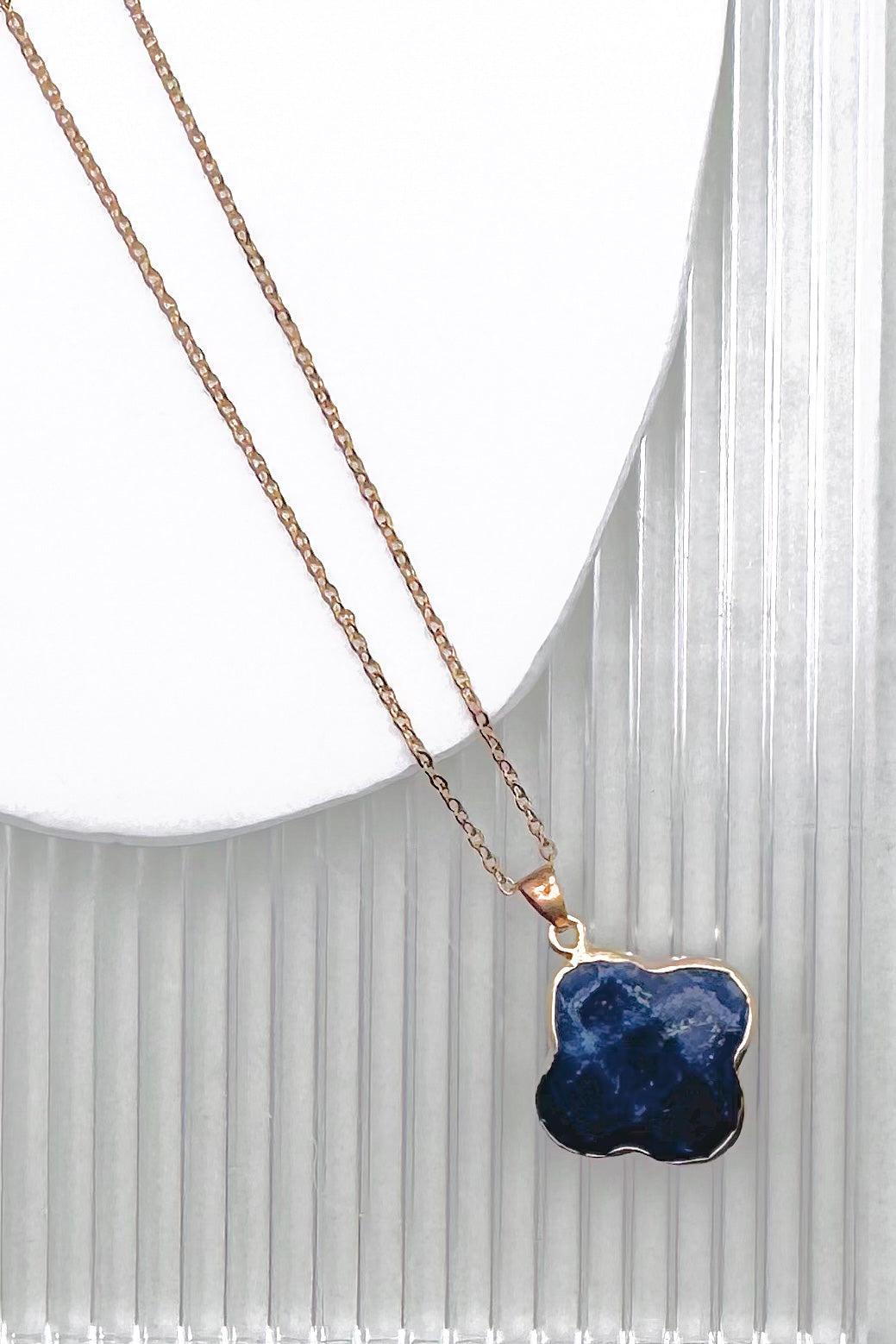 A gold chain necklace with a polished, irregular-shaped dark blue gemstone pendant, displayed on a white curved stand against a white ribbed background.