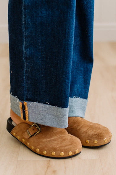 A close-up of a person wearing brown suede slip-on clogs with brass-tone buckles and round metal studs, paired with white socks and dark pants on a light wood floor.