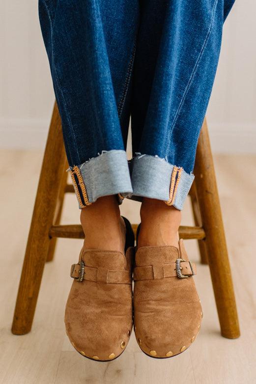 A close-up of a person wearing brown suede slip-on clogs with brass-tone buckles and round metal studs, paired with white socks and dark pants on a light wood floor.
