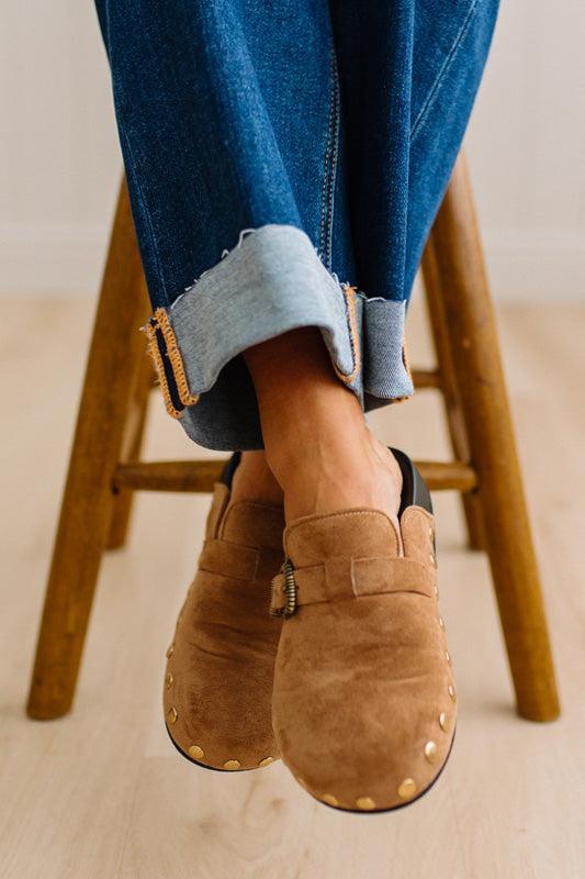 A close-up of a person wearing brown suede slip-on clogs with brass-tone buckles and round metal studs, paired with white socks and dark pants on a light wood floor.