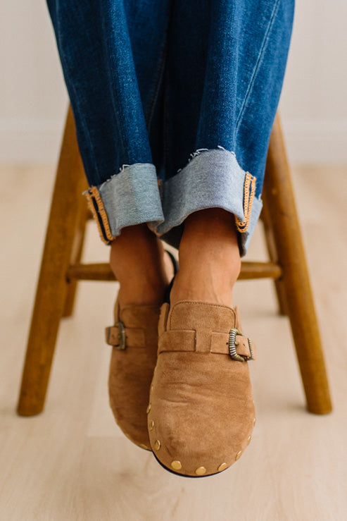 A close-up of a person wearing brown suede slip-on clogs with brass-tone buckles and round metal studs, paired with white socks and dark pants on a light wood floor.