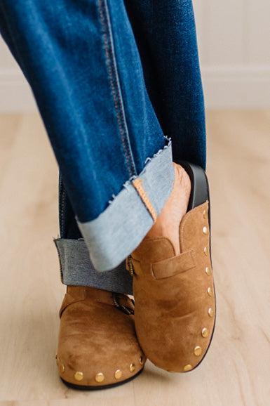 A close-up of a person wearing brown suede slip-on clogs with brass-tone buckles and round metal studs, paired with white socks and dark pants on a light wood floor.