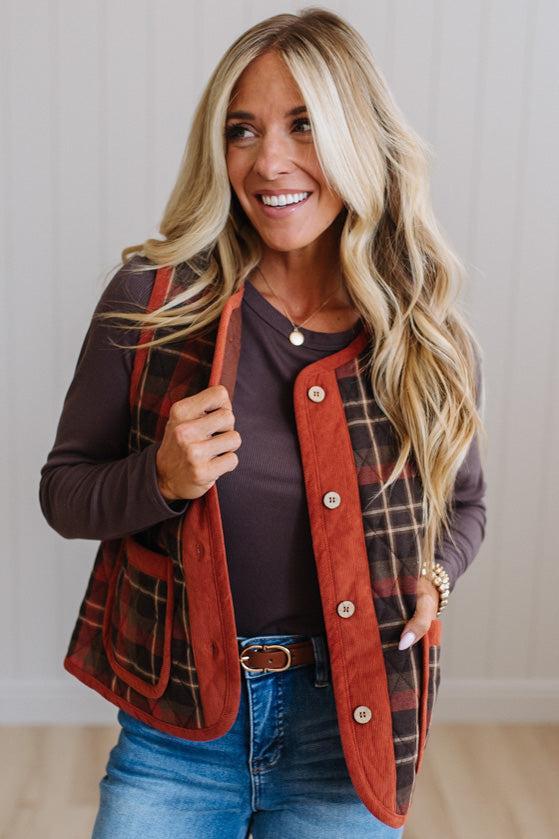 A smiling blonde woman wearing a brown and red plaid button-front vest over a dark brown long-sleeve top and blue jeans, standing against a light paneled wall.