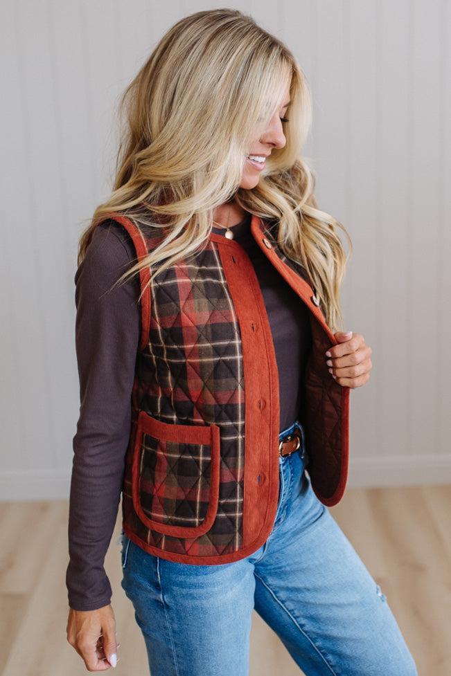 A woman wearing a rust-colored quilted vest with red and black plaid panels, styled over a dark long-sleeve top and blue jeans.