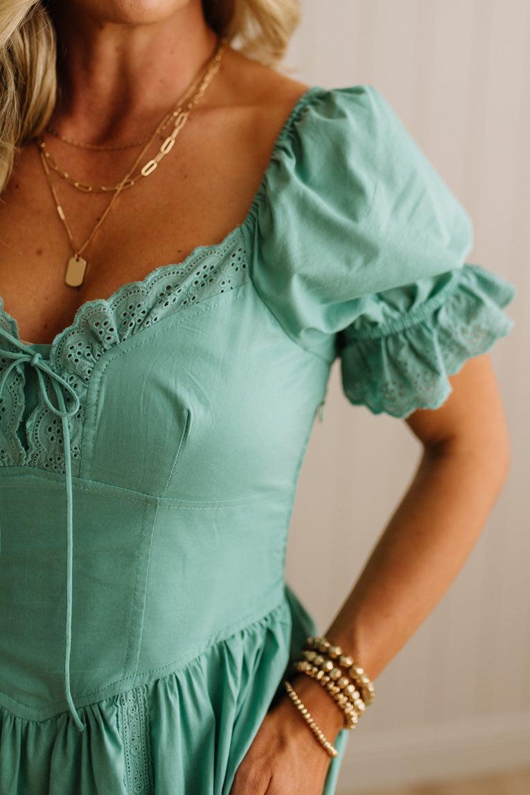 Woman wearing a teal dress with ruffled sleeves and gold jewelry against a neutral background