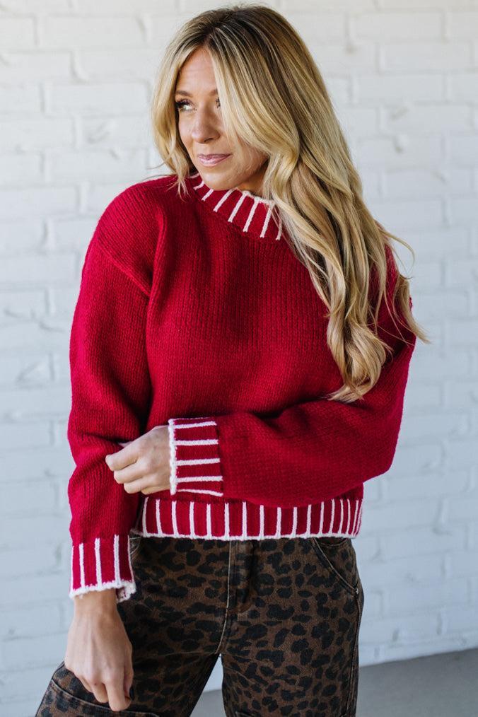 Woman wearing a red sweater with white trim against a white brick wall.