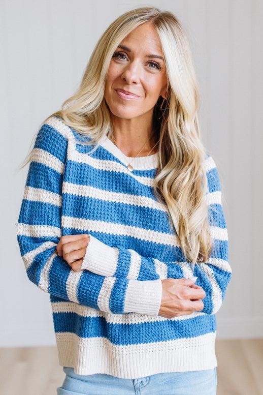 A woman with long blonde hair wearing a blue and white striped knit sweater, standing with her arms crossed against a light gray background.