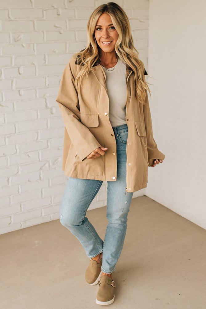 Woman wearing a beige jacket, light blue jeans, and brown shoes against a white brick wall.