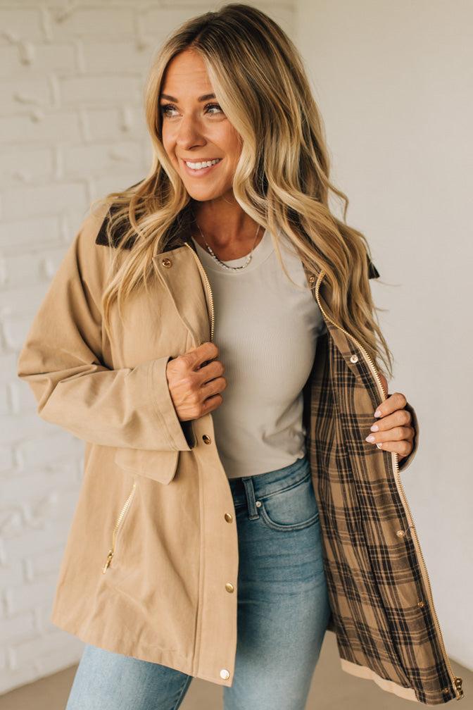 Woman wearing a beige trench coat with plaid lining, white shirt, and blue jeans against a white brick wall.
