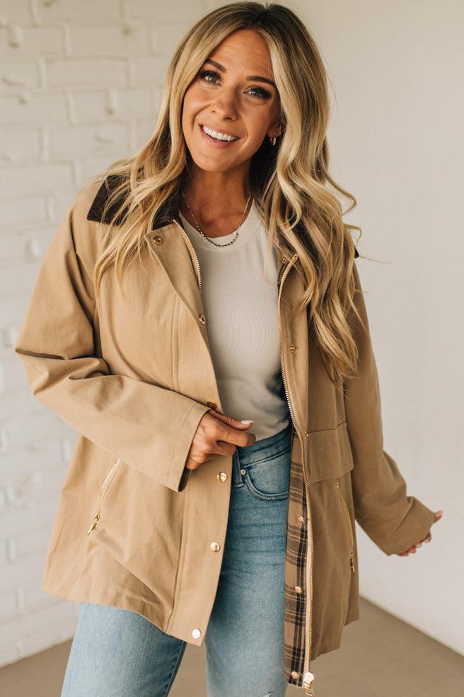 Woman wearing a beige jacket over a white shirt and blue jeans against a white brick wall.