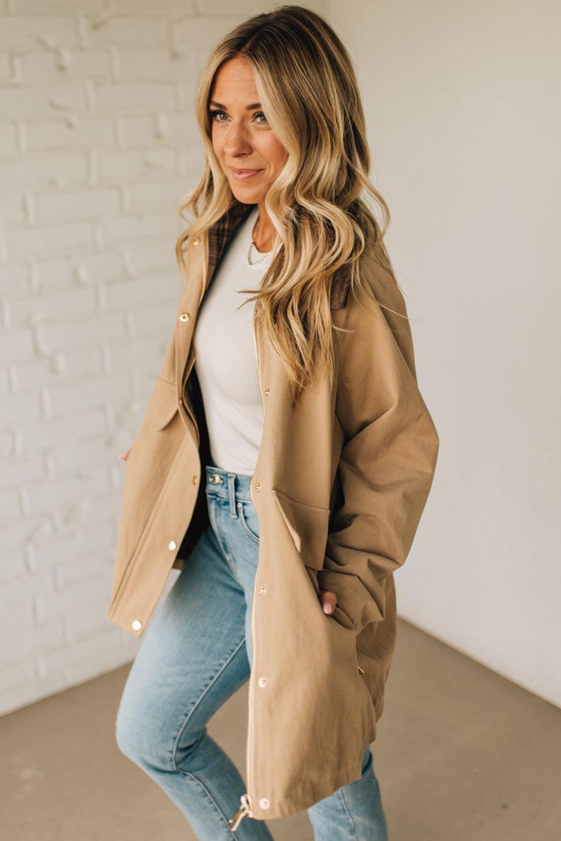 Woman wearing a beige coat over a white shirt and blue jeans against a white brick wall.
