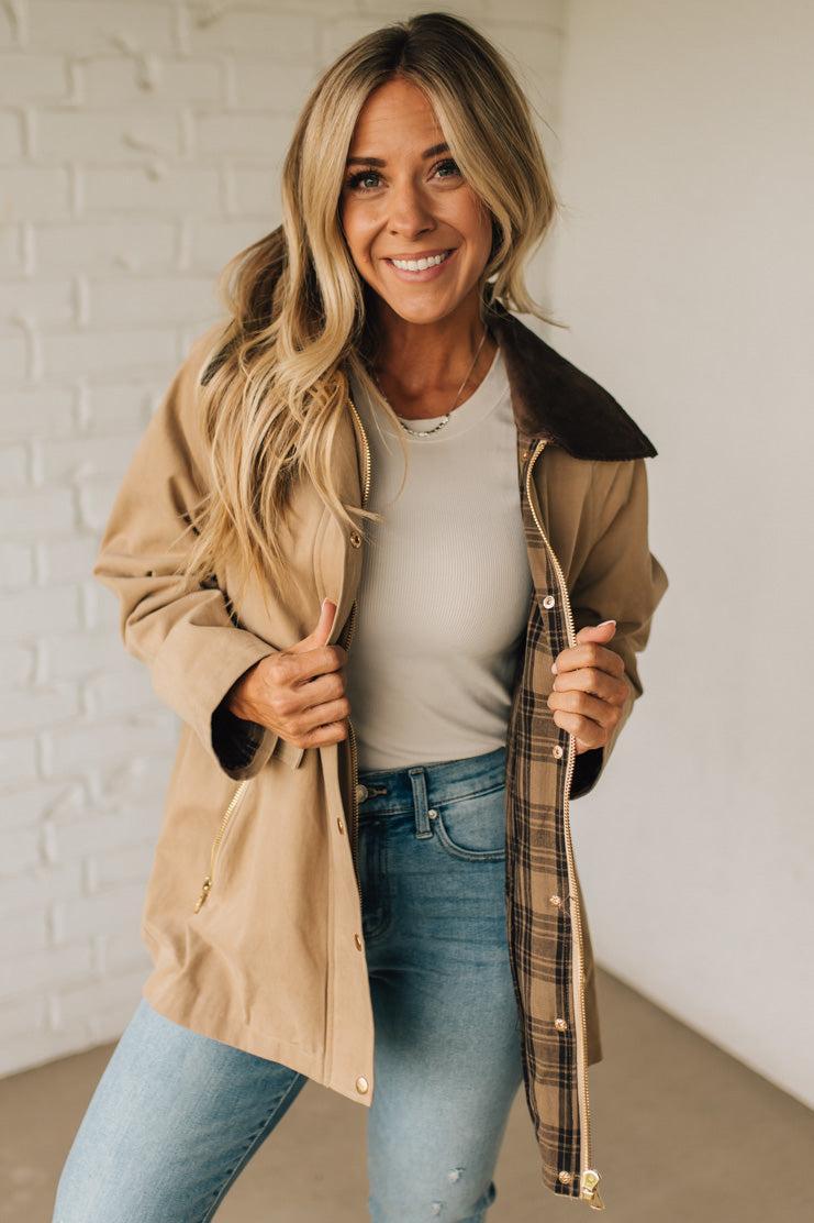 Woman wearing a beige coat with a plaid lining, white shirt, and blue jeans against a white brick wall.
