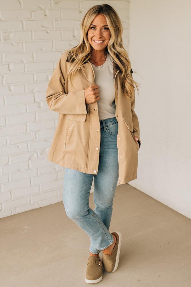 Woman wearing a beige trench coat, white shirt, blue jeans, and brown shoes against a white brick wall.