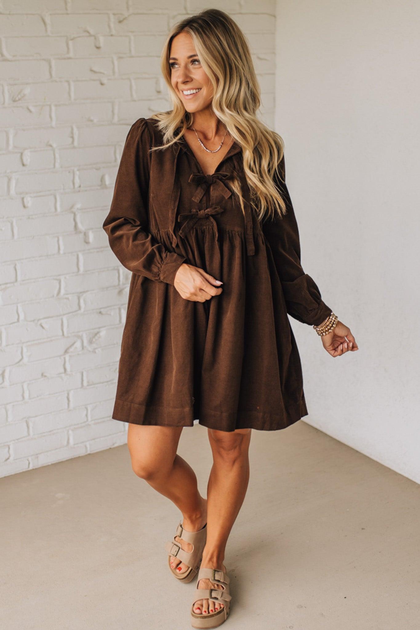 Woman wearing a brown dress standing against a white brick wall.
