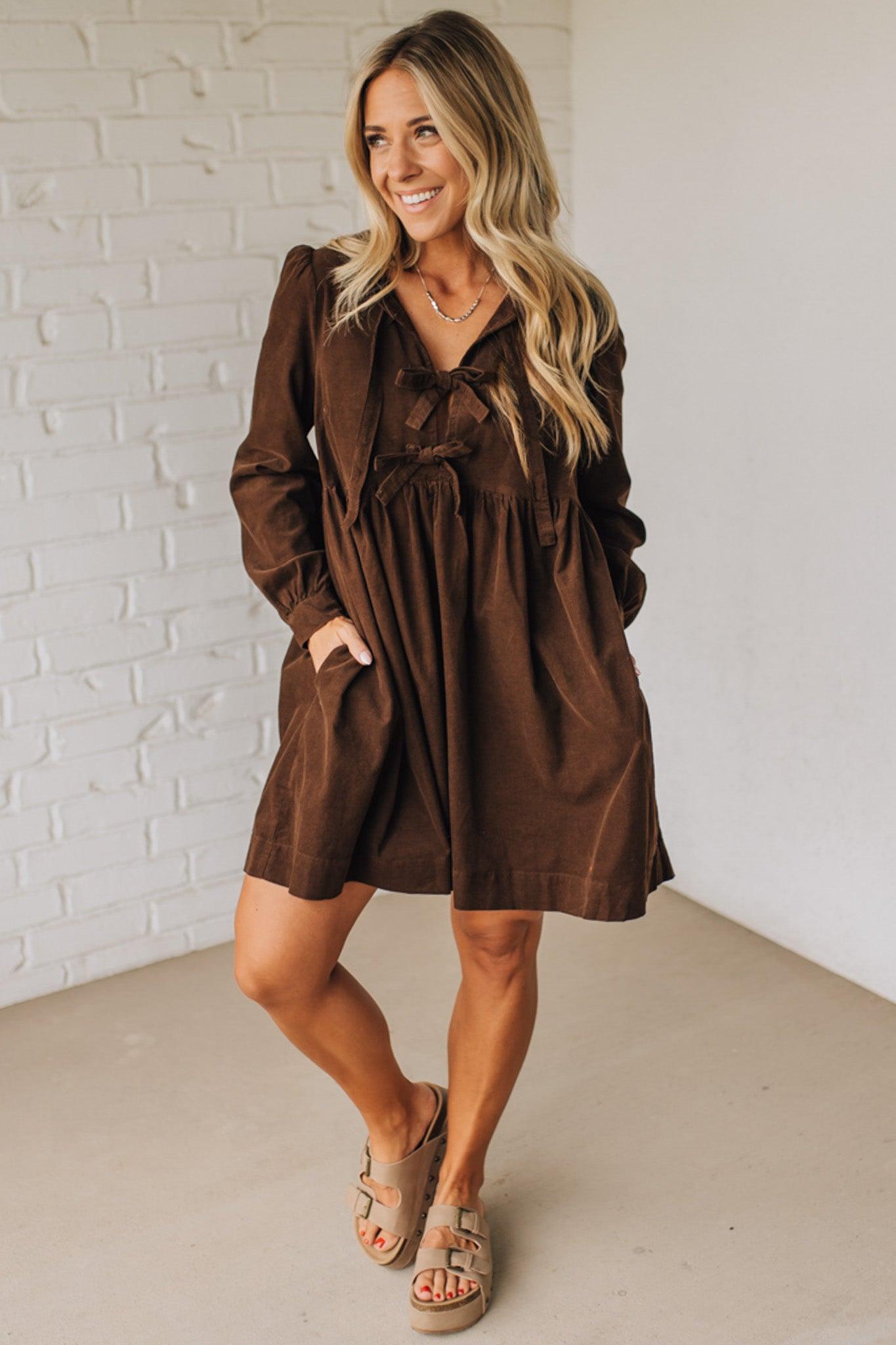 Woman wearing a brown dress standing against a white brick wall.