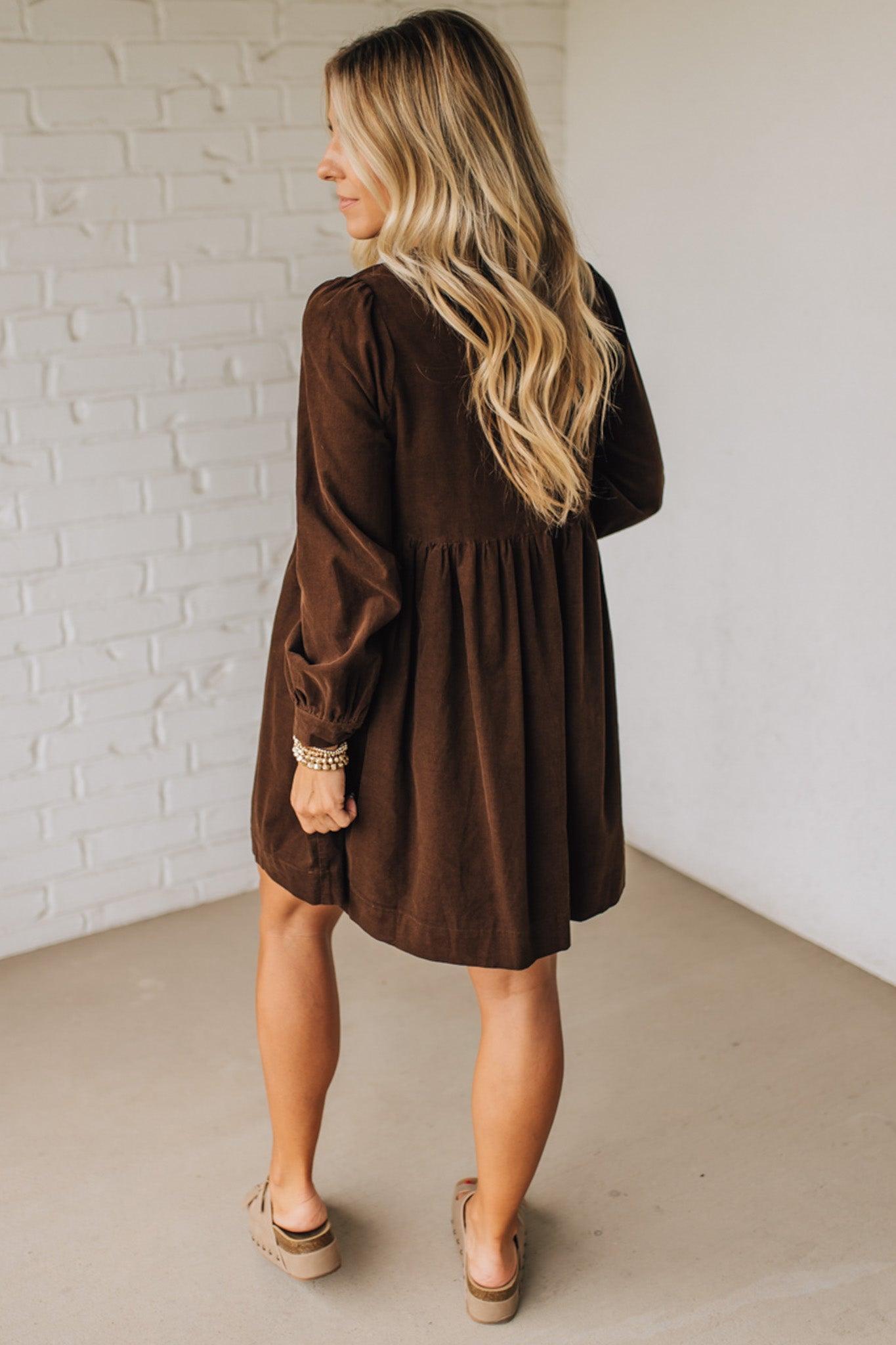 Woman wearing a brown dress standing against a white brick wall.