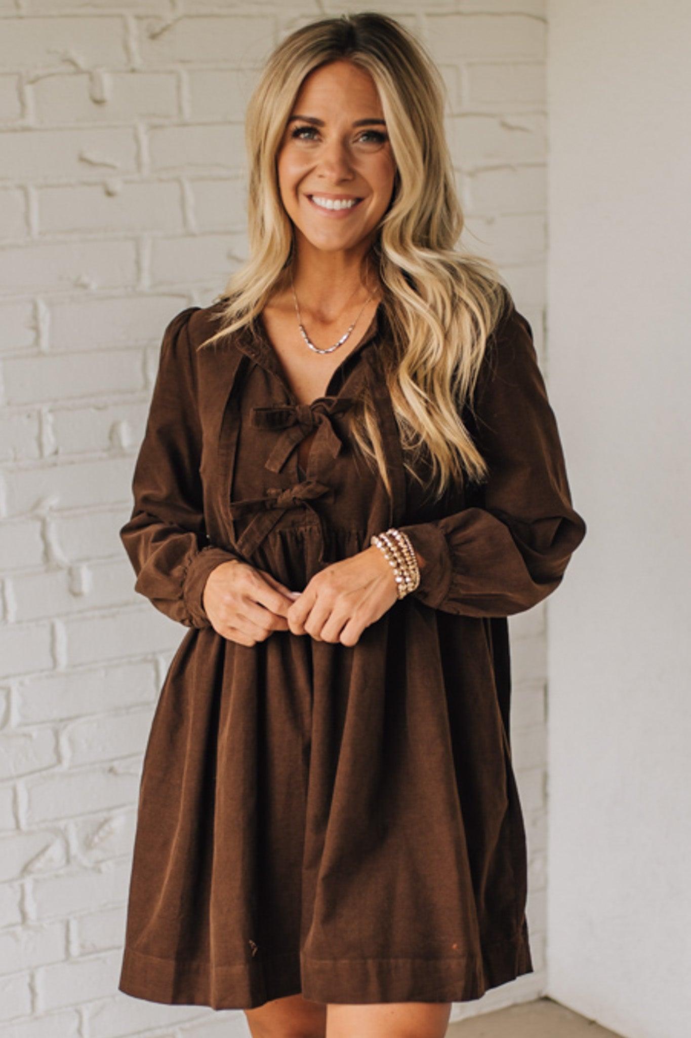 Woman wearing a brown dress standing against a white brick wall.