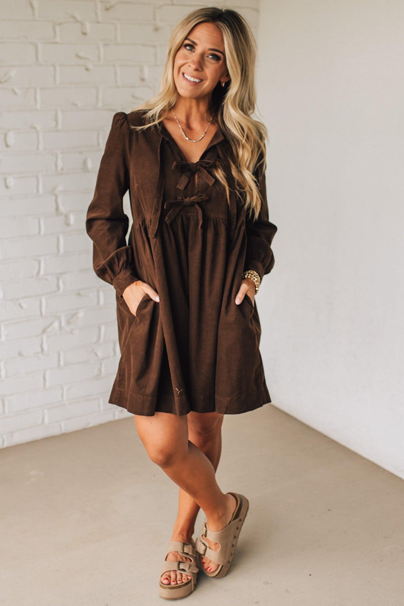Woman wearing a brown dress with a matching jacket against a white brick wall.