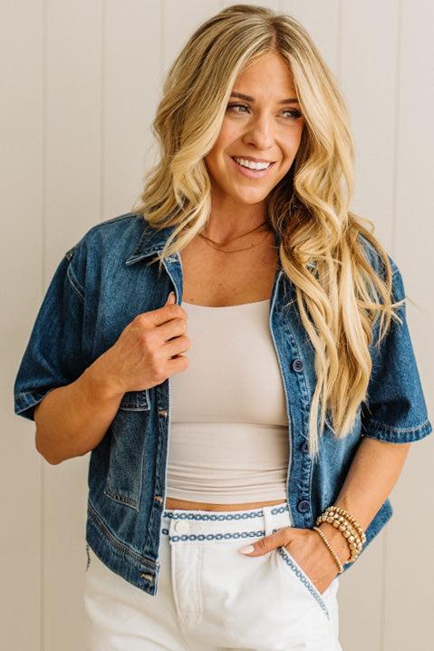 Woman wearing a denim jacket over a white top and pants, smiling against a neutral background