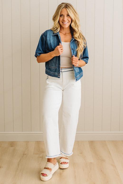 Woman wearing a denim jacket, white pants, and sandals indoors against a light wooden paneled wall.