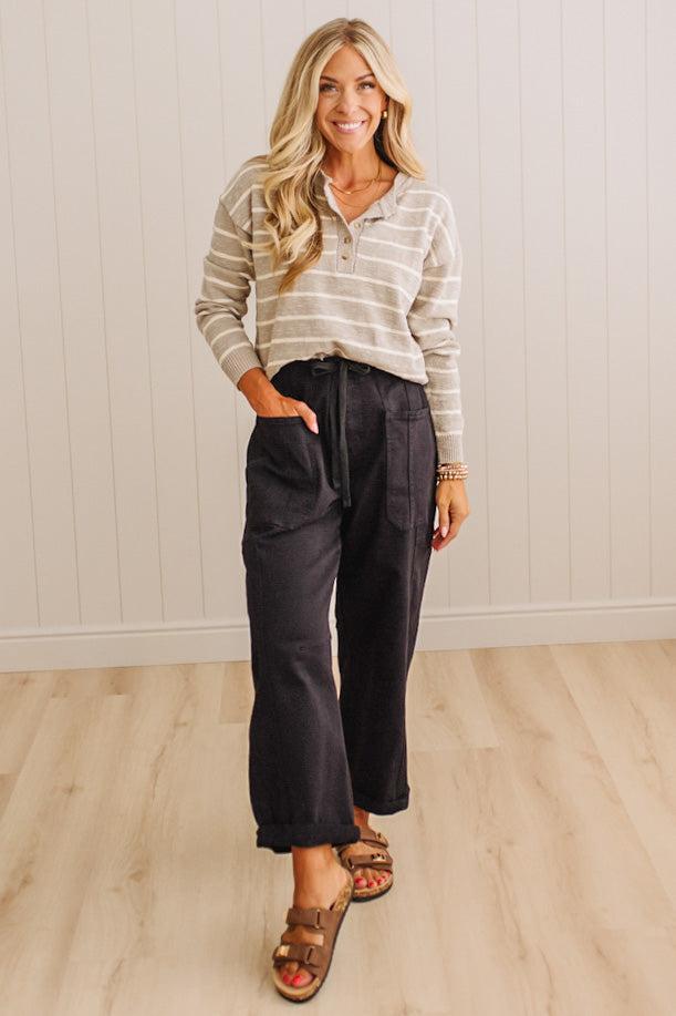 A woman modeling a beige and white striped long-sleeve polo shirt paired with dark charcoal cropped pants and brown leather slide sandals, standing in a room with light wood floors and white paneled walls.