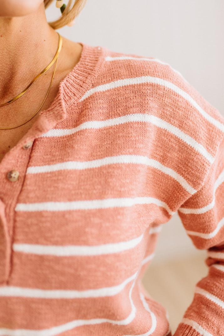 Close-up of a person wearing a soft knit henley sweater with horizontal white stripes on a muted peach-pink base, paired with layered gold necklaces.