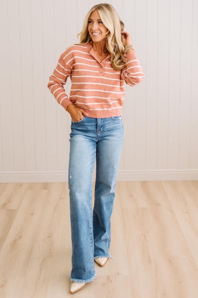 A woman modeling a long-sleeve polo knit top with horizontal peach and white stripes, paired with light-wash blue jeans and beige pointed-toe flats, standing in a brightly lit room with light wood floors and white paneled walls.