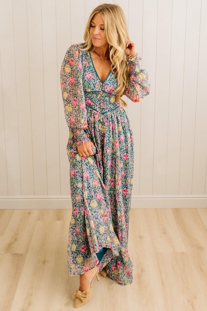 Woman wearing a floral dress standing in a room with wooden flooring and a white wall.