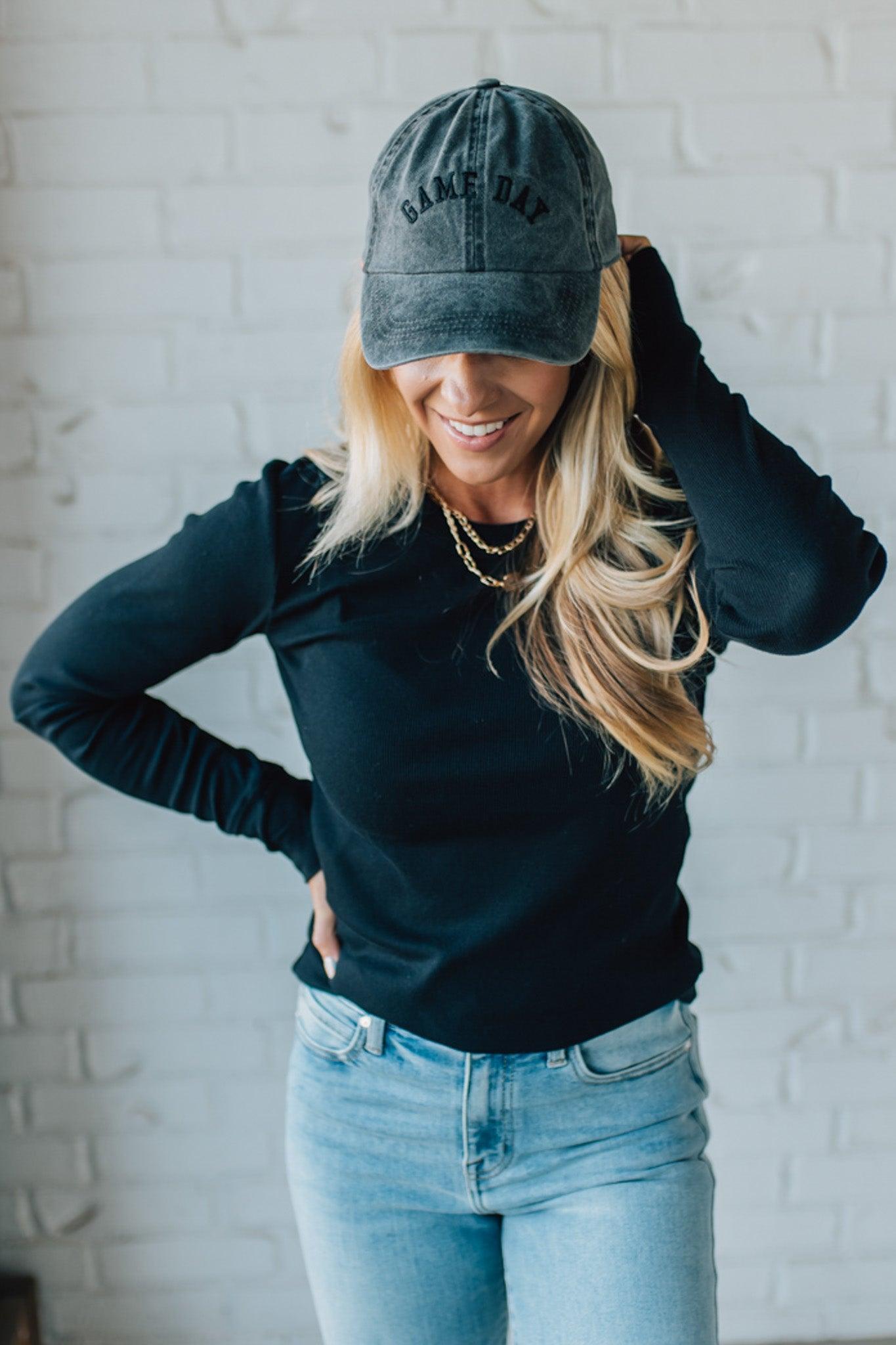 Woman wearing a black long-sleeve top, light blue jeans, and a gray cap with 'Bake Day' text against a white brick wall.