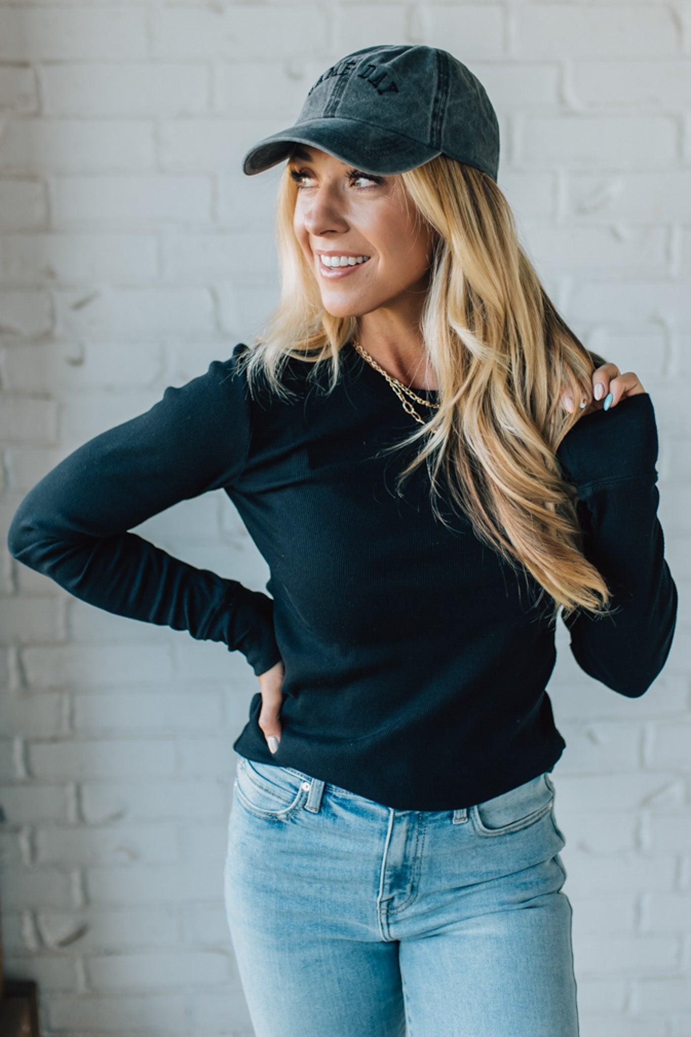 Woman wearing a black long-sleeve top, light blue jeans, and a dark baseball cap against a white brick wall.