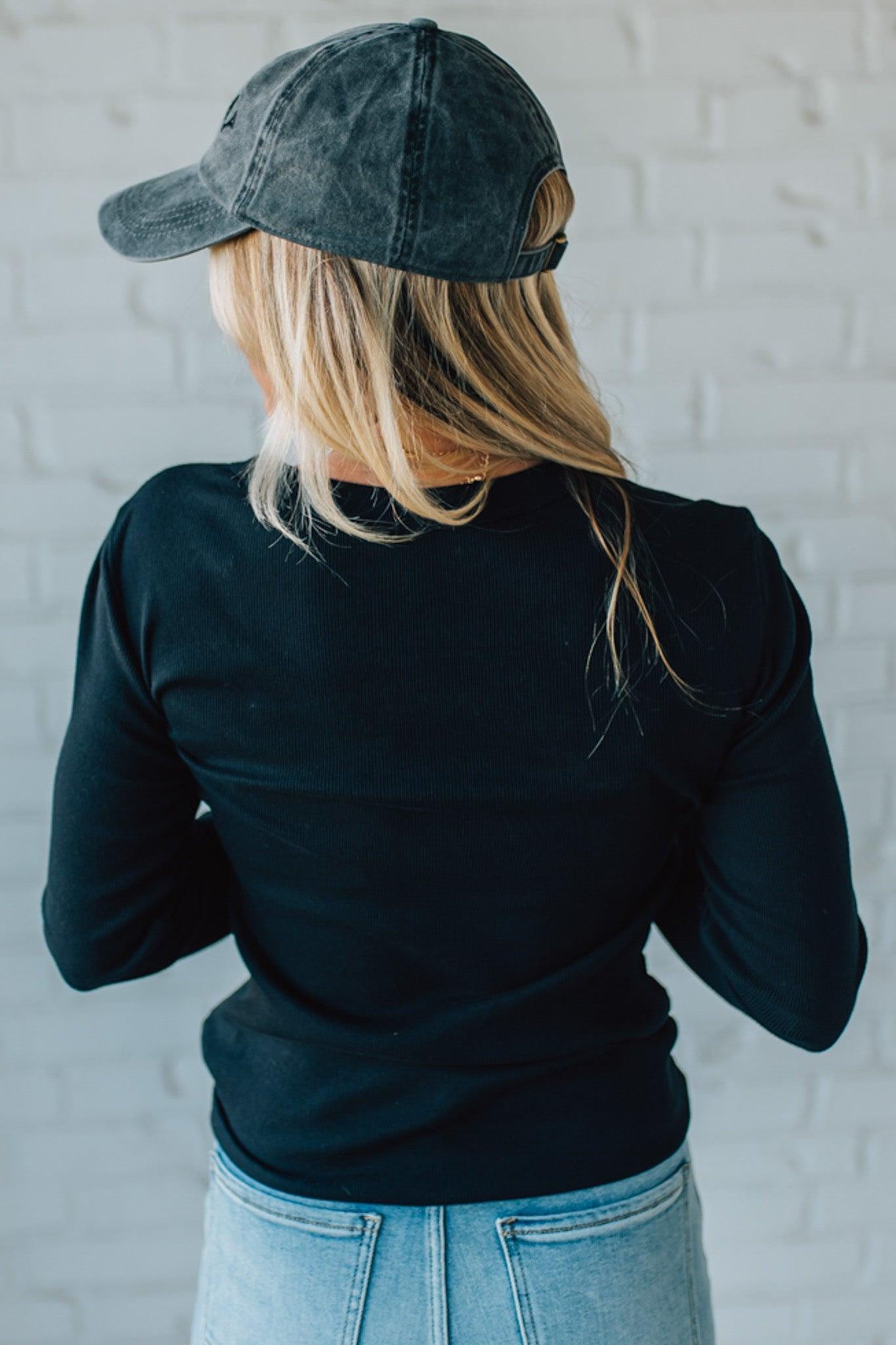 Person wearing a black long-sleeve shirt and blue jeans with a gray cap, standing against a white brick wall.